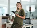 Uśmiechnięta ciężarna kobieta z tabletem i kubkiem w biurze. Dowiedziałam się o ciąży po okresie wypowiedzenia, ale teraz planuję przyszłość.
