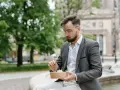 Mężczyzna w okularach i marynarce robi sobie przerwę w pracy, jedząc sałatkę na ławce w parku.