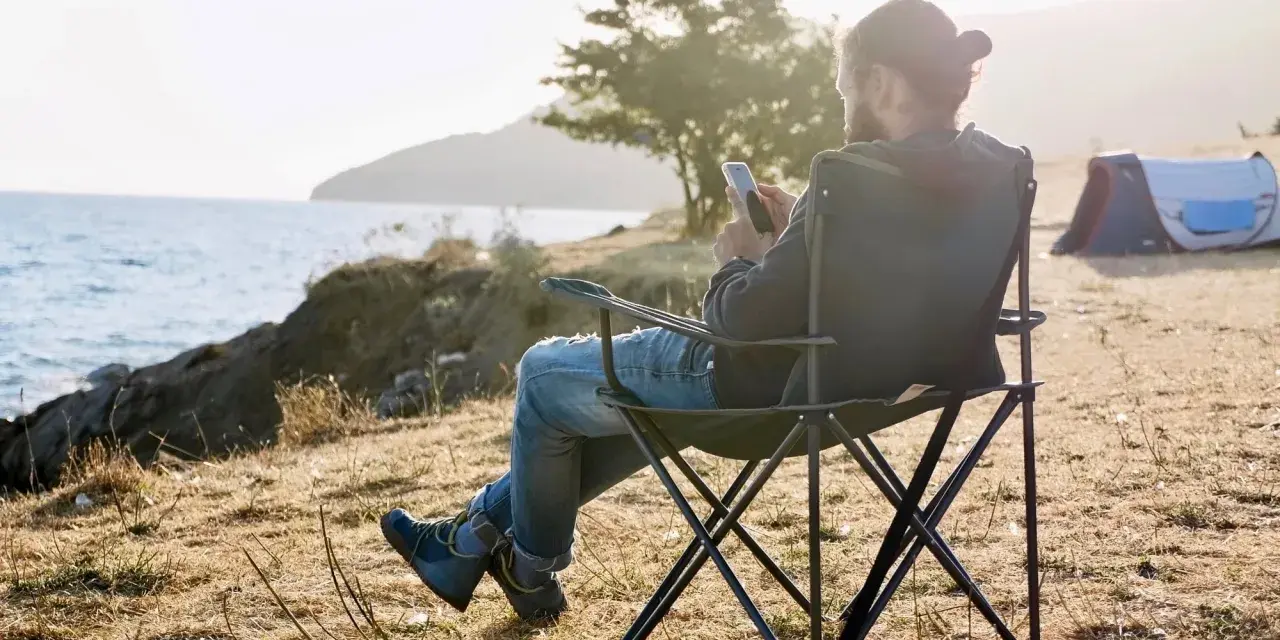 Mężczyzna na urlopie bezpłatnym siedzi na krześle kempingowym nad morzem, sprawdzając telefon. Obok namiot.