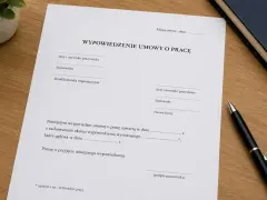 Dokument "Wypowiedzenie umowy o pracę z zachowaniem okresu wypowiedzenia" na drewnianym biurku, obok długopisu i książki.