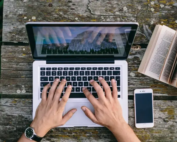Ręce piszą CV funkcjonalne wzór na laptopie. Obok leży otwarta książka i telefon.