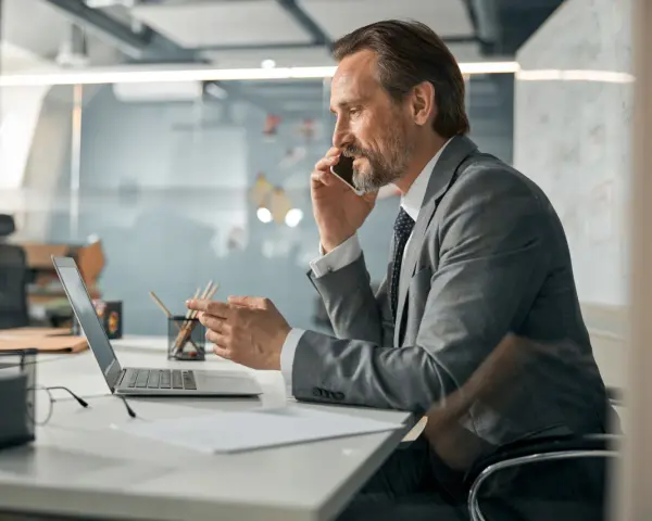 Biznesmen w garniturze rozmawia przez telefon, pracując przy laptopie. To typowy dzień w życiu CEO, co to znaczy być liderem.