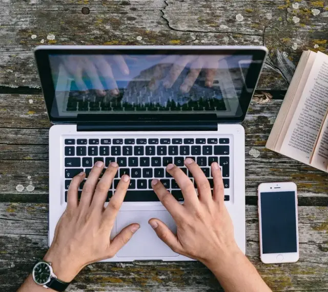 Ręce piszą CV funkcjonalne wzór na laptopie. Obok leży otwarta książka i telefon.